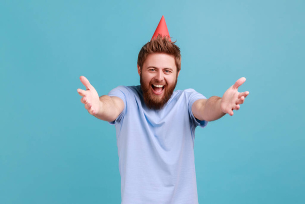 Portrait of extremely happy bearded man wearing party cone giving free hugs with outstretched hands, welcoming inviting to embrace. Indoor studio shot isolated on blue background. - Foto, Imagen