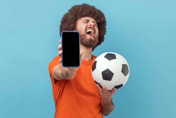 Extremely happy man with Afro hairstyle wearing T-shirt standing with soccer ball and showing mobile phone with empty display, betting and winning. Indoor studio shot isolated on blue background. - Photo, image