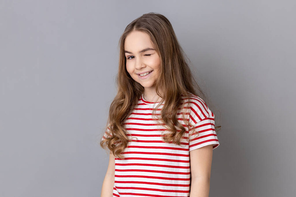 Portrait of playful cheerful little girl wearing striped T-shirt standing and winking playfully, positive expression, looking at camera. Indoor studio shot isolated on gray background. - Photo, Image