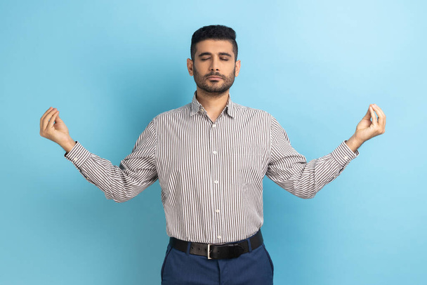 Portrait of calm relaxed young businessman with beard standing with raised arms and doing yoga meditating exercise, wearing striped shirt. Indoor studio shot isolated on blue background. - Zdjęcie, obraz