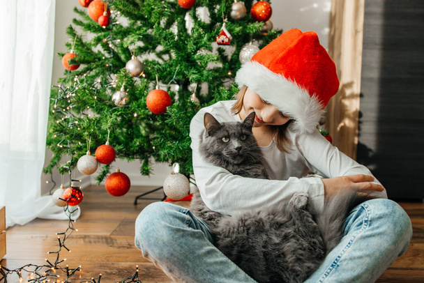 サンタの帽子の女の子は、クリスマスツリーの隣のクリスマスの前夜に灰色の猫を抱きかかえています。面白いペット. - 写真・画像
