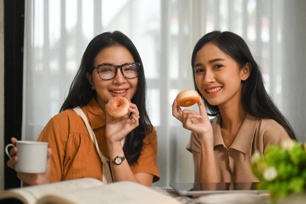 Due felici giovani asiatiche donne migliori amici mangiare ciambelle e godersi il tempo libero insieme a casa. - Foto, immagini