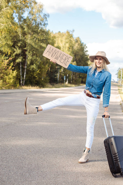 Vertical begging, funny, smiling blond woman autostopping, hitchhiking by raised leg sign, with cardboard plate anywhere carrying luggage on road. Take anywhere, new life adventure and travelling - Photo, Image