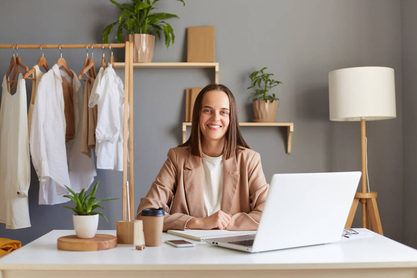 Portrait of happy satisfied woman designer working on laptop in living room against clothes hang on shelf, sitting at table and looking at camera with toothy smile, works in her showroom. - 写真・画像