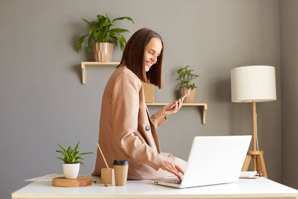 Indoor shot of young adult adorable happy businesswoman wearing jacket working on home office and using laptop and mobile phone, being in good mood at work. - Zdjęcie, obraz