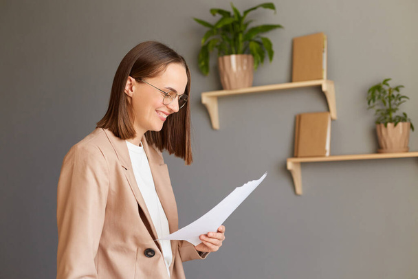 Indoor shot of Caucasian business woman holding holding paper documents, working in office, wearing beige jacket, posing against gray walls in office. - Фото, изображение