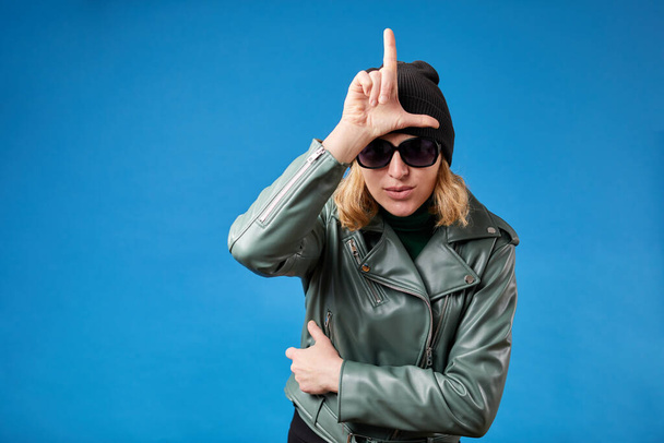 Portrait of young woman with blonde hair in green jacket standing, showing loser gesture with hand on forehead. - Foto, imagen