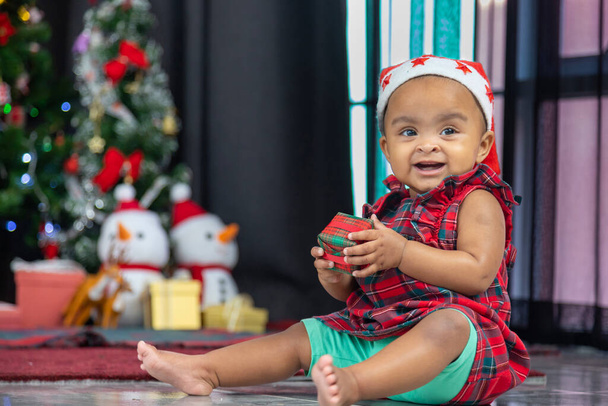 African American baby is happily smiling while receiving little gift box from parents while dressing in christmas dress and santa hat with christmas tree on the back for season celebration  - Фото, изображение