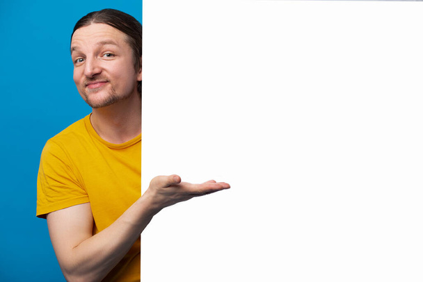 Grappige man geeft vinger poster lege ruimte, dragen oranje shirt geïsoleerde blauwe kleur achtergrond. Korting, verkoop concept - Foto, afbeelding