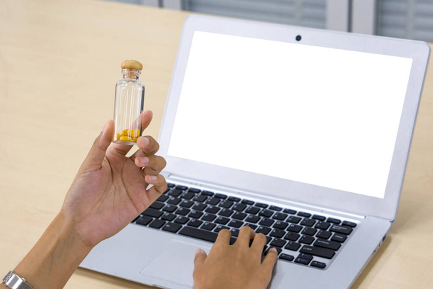 Closeup hand holding glass pill packer bottle with wooden cap in front of white screen laptop computer. - Photo, Image