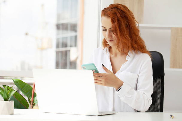 Red-Haired Woman Talking On Mobile Phone Working Remotely On Laptop. - 写真・画像