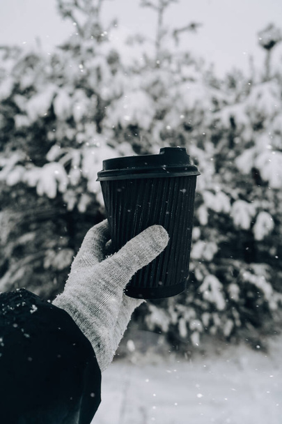 Female hand in winter warm gloves holding black eco paper cup. Winter picnic outdoors. Creative trendy zero waste recycle cup. Mockup. Hot warming drink to go in winter day. Take away concept leisure concept tea or coffee cup - Foto, Imagen
