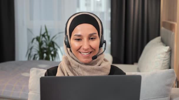 Csinos fiatal arab muszlim nő blogger fülhallgató ül otthon irodai laptopot, amelynek video online hívás. Női vlogger hidzsábban dolgozik, távolsági kurzusok. Videó blogolás. Muszlim. Oktatás - Felvétel, videó