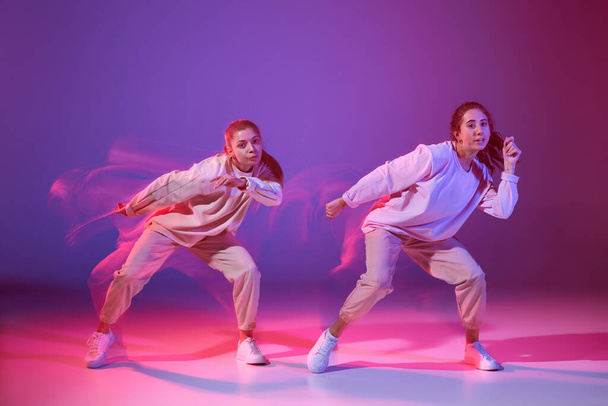 Portret dwóch młodych dziewcząt tańczących hip-hop odizolowany od gradientowego fioletowego tła neonem ze światłem mieszanym. Pojęcie ruchu, kultura młodzieżowa, aktywny styl życia, akcja, taniec uliczny - Zdjęcie, obraz