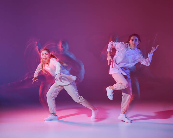 Retrato de chicas jóvenes bailando hip-hop aisladas sobre fondo violeta degradado en neón con luz mixta. Coge el ritmo. Concepto de movimiento, cultura juvenil, estilo de vida activo, acción, danza callejera - Foto, Imagen