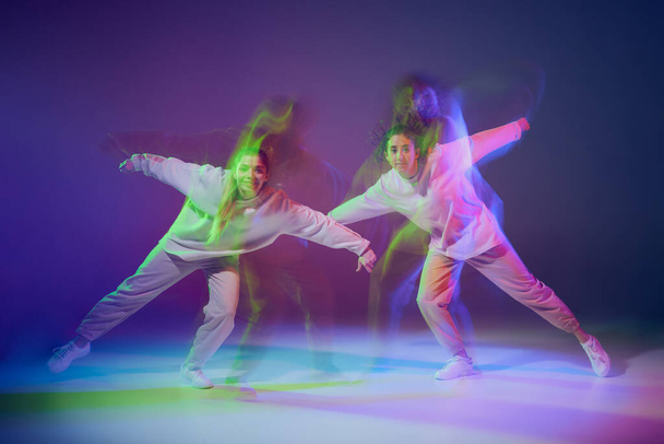 Портрет молодих дівчат, що танцюють хіп-хоп, ізольований на градієнтному синьо-фіолетовому фоні в неоні зі змішаним світлом. Дружба. Концепція руху, молодіжна культура, активний спосіб життя, дії, вуличний танець
 - Фото, зображення