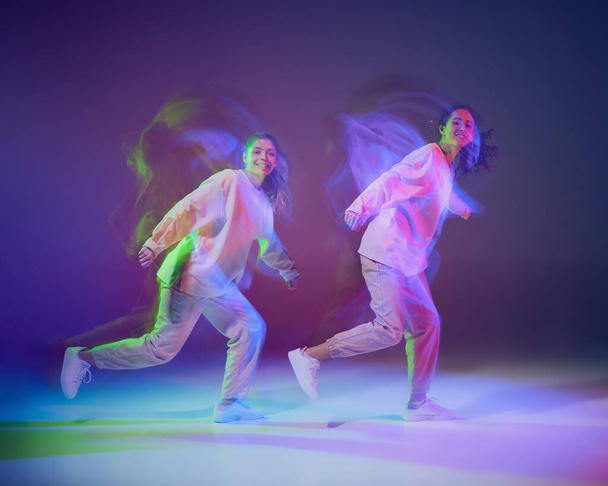 Портрет молодых девушек, танцующих хип-хоп, выделен на градиентно-синем фиолетовом фоне в неоновом с смешанным светом. Выглядишь счастливым. движение, молодежная культура, активный образ жизни, экшн, уличные танцы - Фото, изображение