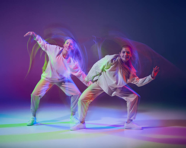 Young girls dancing hip-hop, performing on stage isolated over gradient blue purple background in neon with mixed light. Concept of movement, youth culture, active lifestyle, action, street dance - Foto, Imagem