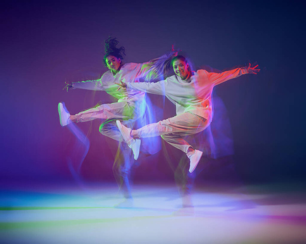 Portrait of young girls dancing hip-hop isolated over gradient blue purple background in neon with mixed light. Jump. Concept of movement, youth culture, active lifestyle, action, street dance - Photo, Image