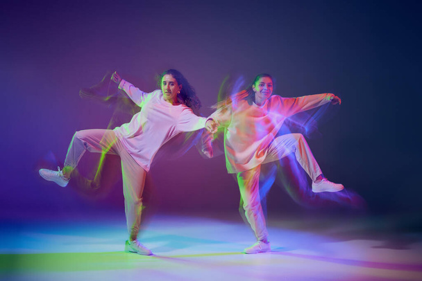 Portrét mladých dívek tančících hip-hop izolované na gradient modré purpurové pozadí v neonu se smíšeným světlem. Zábava. Koncepce pohybu, kultura mládeže, aktivní životní styl, akce, pouliční tanec - Fotografie, Obrázek
