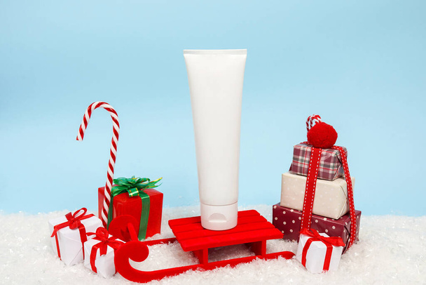 Tubo de crema cosmética permaneciendo en trineo rojo cerca de cajas de regalo de Navidad sobre fondo azul. Venta de Navidad y Año Nuevo, regalos, belleza y cuidado de la piel concepto - Foto, imagen