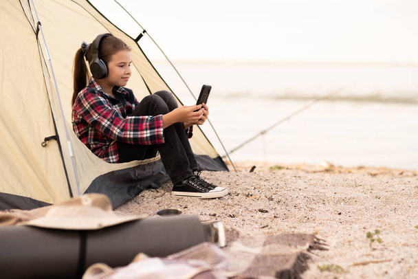 Young tourist on vacation in seashore. Happy teen girl relaxing in camping tent. Hiker child listen to music from the phone. People living healthy active lifestyle. Copy space - Photo, Image
