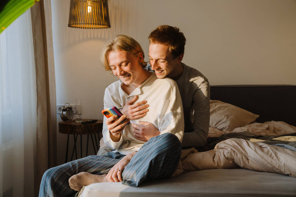 Young gay couple hugging and using cellphone while sitting in bed at home - Foto, Imagem
