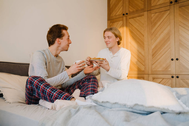 Jeune couple gay souriant et prenant le petit déjeuner tout en se reposant au lit à la maison - Photo, image
