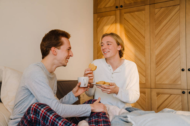 Young gay couple smiling and having breakfast while resting in bed at home - Foto, imagen