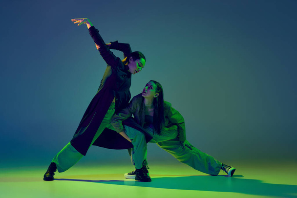 Emotions. Stylish young girls in casual clothes dancing experimental style dance over gradient blue-green background at dance hall in neon light. Youth culture, contemporary dance, fashion, action. - Foto, Imagem