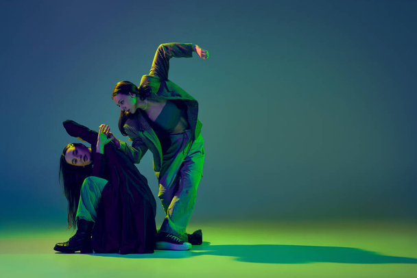 Young stylish flexible girls in modern attires dancing contemp style dance isolated over gradient blue-green background at dance hall in neon light. Art, contemporary dance, fashion, action, ad - Foto, immagini