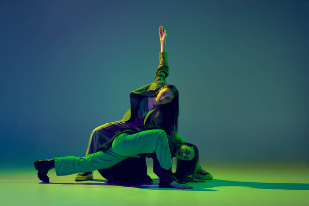 Stylish young girls in casual clothes dancing experimental style dance over gradient blue-green background at dance hall in neon light. Youth culture, contemporary dance, fashion, action. - Foto, Imagen