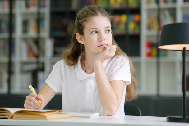 School girl wearing white t-shirt sitting at table and writing in exercise book, looking to side and daydreaming, bookshelves on blurred background. Pupil doing assignment in class - Photo, image