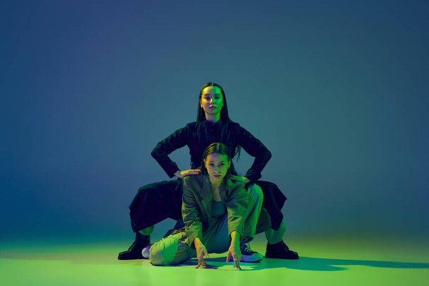Young stylish flexible girls in modern attires dancing contemp style dance isolated over gradient blue-green background at dance hall in neon light. Art, contemporary dance, fashion, action, ad - Photo, Image