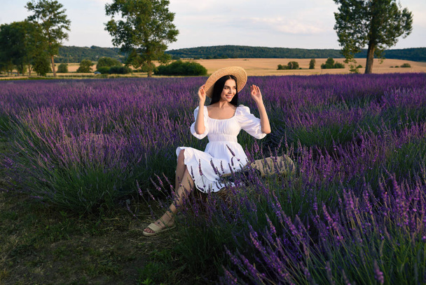 Beautiful young woman sitting in lavender field - 写真・画像