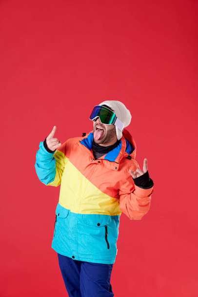 Young boy in cap, jacket and snow goggles sticking out his tongue and holding up the fingers of both hands on a red background. - Fotó, kép