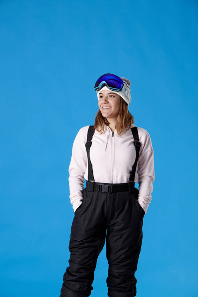 glimlachend meisje kijkt naar rechts met sneeuw gear, hoed en bril op het hoofd op een blauwe achtergrond. - Foto, afbeelding