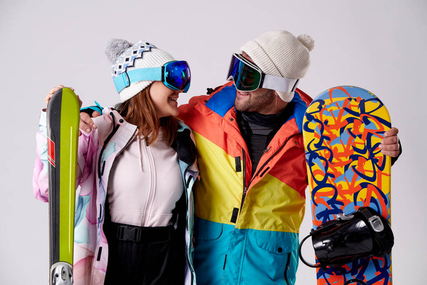 boy and girl looking at each other smiling with snow goggles holding a snowboard and some skis on a white background. - Photo, Image