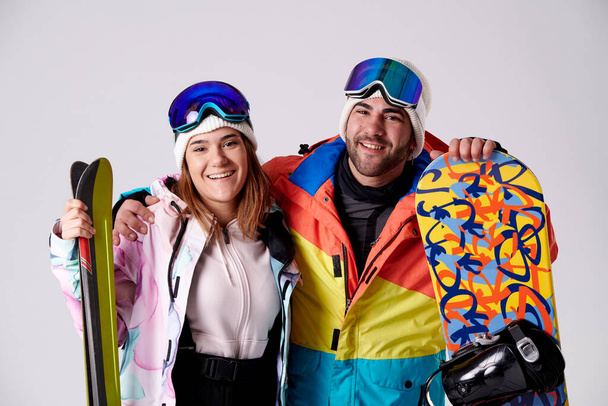 uśmiechnięty chłopiec i dziewczyna trzymając snowboard i narty patrząc w aparat na pierwszym planie na białym tle. - Zdjęcie, obraz