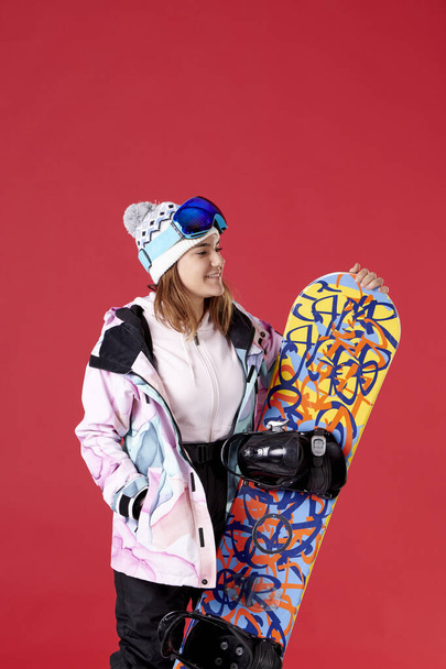 girl wearing winter clothes, hat and snow goggles holding a snowboard on a red background. - Fotó, kép