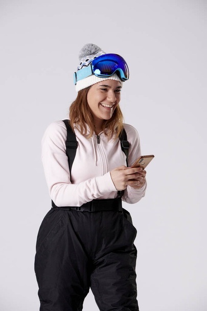 Девушка в шляпе, очках и снегоходах печатает на мобильном телефоне, улыбаясь на белом фоне. - Фото, изображение