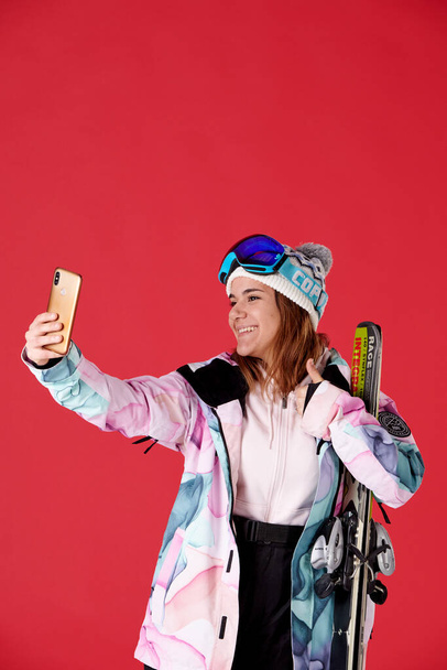 Девушка в снежном снаряжении держит пару лыж, делая селфи, улыбаясь на красном фоне. - Фото, изображение