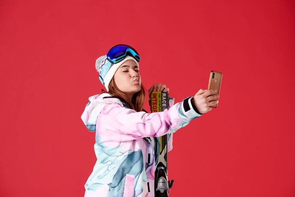 lány sílécek és hó felszerelés nézi a mobiltelefon csinál egy szelfi kacsintás és ad egy csókot a piros háttér. - Fotó, kép