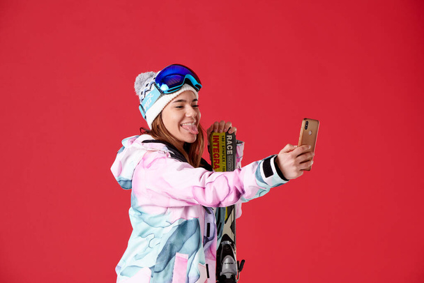 lány sílécek és hó felszerelés nézi a mobiltelefon csinál egy szelfi a nyelve ki a piros háttér. - Fotó, kép