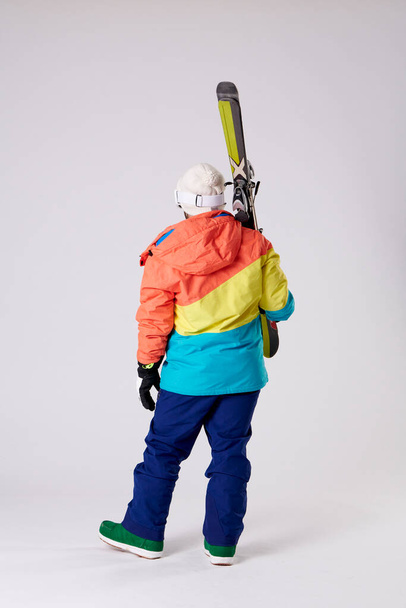 chłopiec z tyłu z odzieżą śnieżną trzymając narty jedną ręką na białym tle. - Zdjęcie, obraz