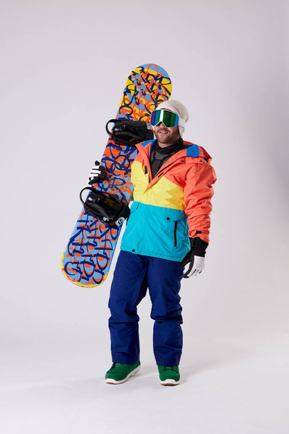 Şapkalı, gözlüklü, kar gözlüklü bir çocuk bir eli beyaz bir arka planda olan bir snowboard tutuyor.. - Fotoğraf, Görsel
