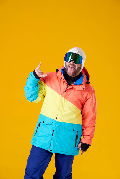 Junge in Mütze, Winterkleidung und Snowboardbrille streckt die Zunge in die Kamera und macht die Hörner mit den Fingern seiner Hand auf gelbem Hintergrund. - Foto, Bild