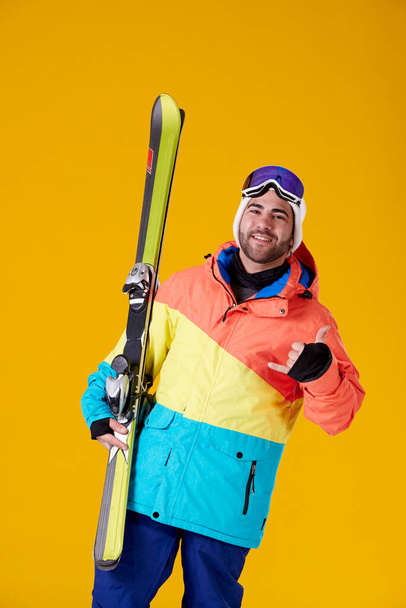 Lächelnder junger Mann in Snowboardkleidung hält ein paar Skier in der Hand und macht das Good-Vibes-Symbol mit seiner Hand, die in die Kamera auf gelbem Hintergrund blickt. - Foto, Bild