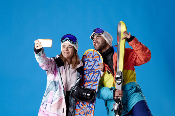 nainen ja mies ottaa selfie lumessa vaihde osoittaa niiden sukset ja lumilaudat sinisellä pohjalla. - Valokuva, kuva