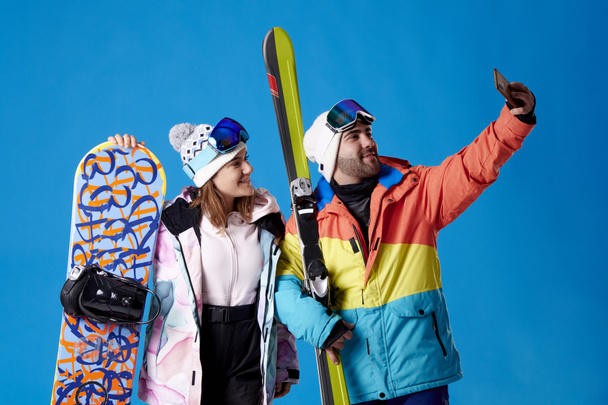 young man and woman on skis and snowboard taking a selfie while smiling on a blue background. - Zdjęcie, obraz